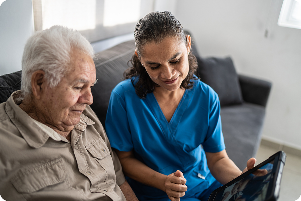 A healthcare professional in blue scrubs engages with an elderly man while displaying content on a tablet.