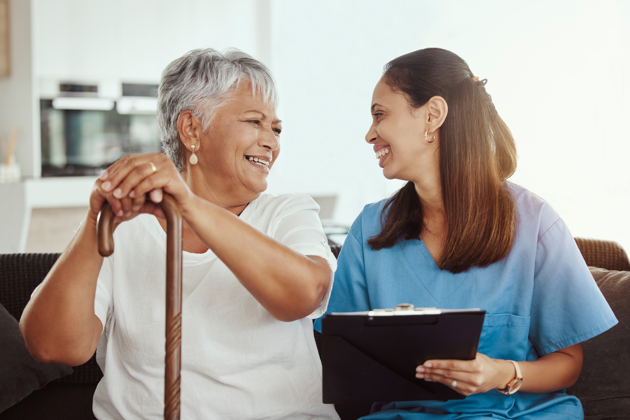 A smiling elderly woman with a cane interacts warmly with a healthcare professional holding a clipboard in a cozy setting.