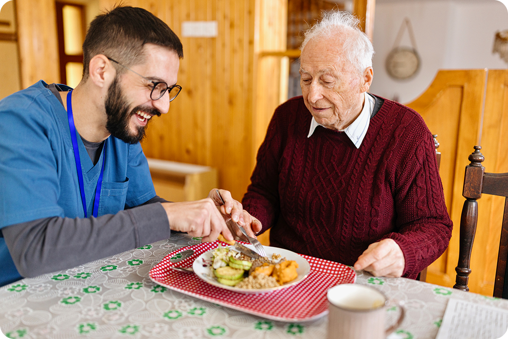 Caregiver assisting an elderly man with a meal at a table, featuring a plate of food and a warm, inviting interior.