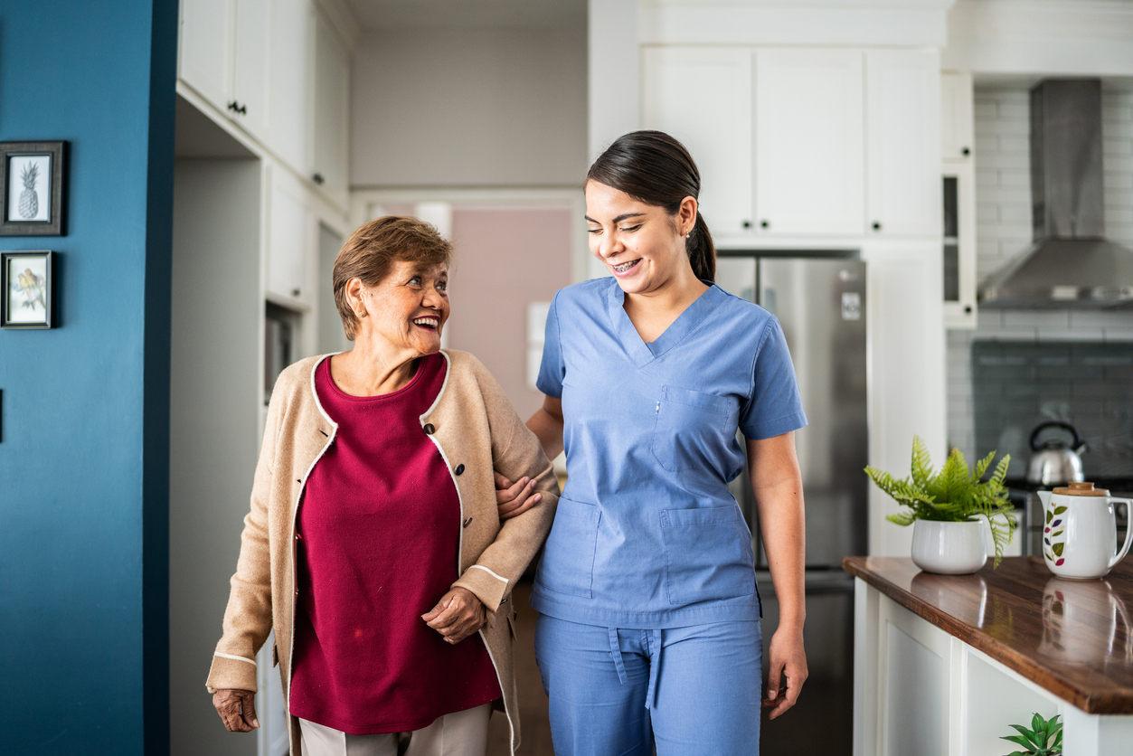 A healthcare worker assists an elderly woman, both smiling, in a bright, welcoming kitchen environment.