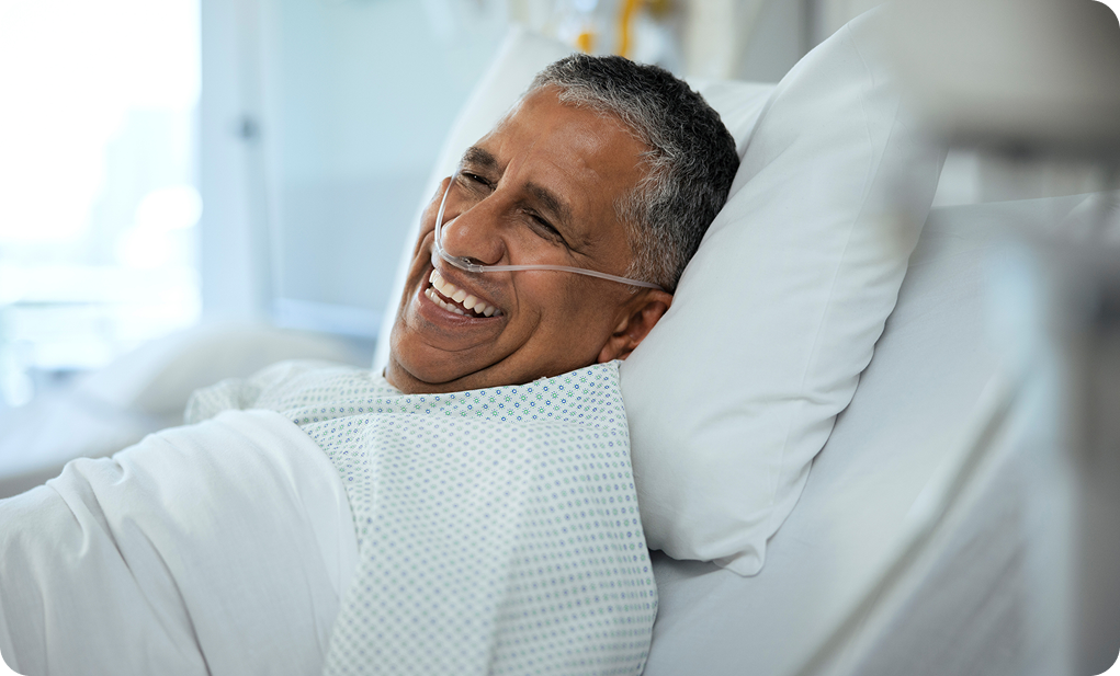 A smiling patient wearing a hospital gown and nasal cannula, comfortably resting in a bright hospital room.
