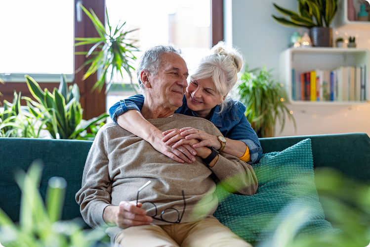 An older man and woman share a warm embrace on a couch, surrounded by houseplants and a cozy living room setting.