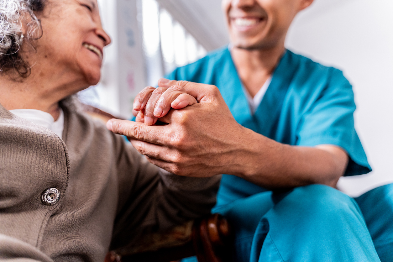 A caregiver and a senior woman share a compassionate moment, clasping hands in a well-lit, comfortable environment.