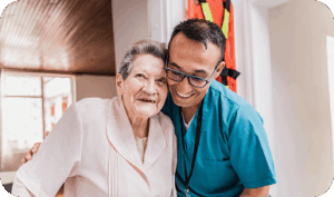 A smiling healthcare professional embraces an elderly woman in a brightly lit indoor setting, conveying warmth and care.