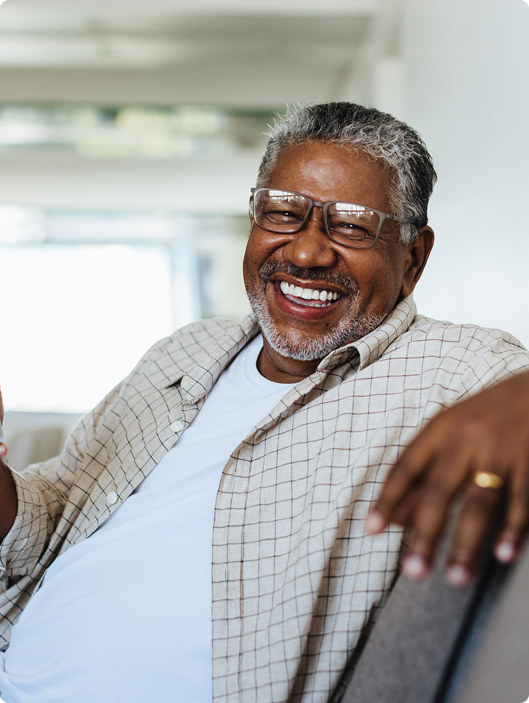 Smiling elderly man with glasses and gray hair relaxing in a bright indoor setting, wearing a plaid shirt over a white shirt.