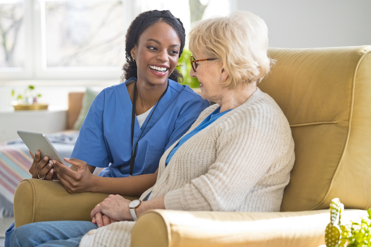 Nurse in blue scrubs engages with an elderly woman while showing her a tablet in a bright, comfortable room.