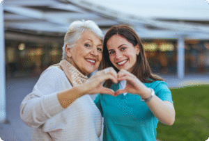 Portrait of a smiling elderly woman and a nurse making a heart shape with their hands in a welcoming outdoor setting.