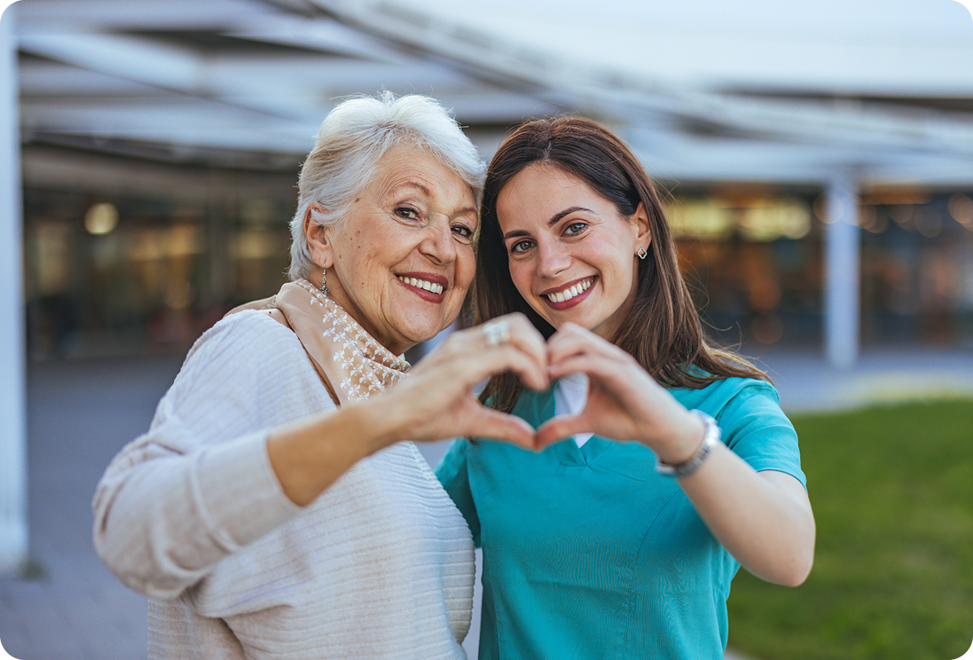 Portrait of a smiling elderly woman and a nurse making a heart shape with their hands in a welcoming outdoor setting.
