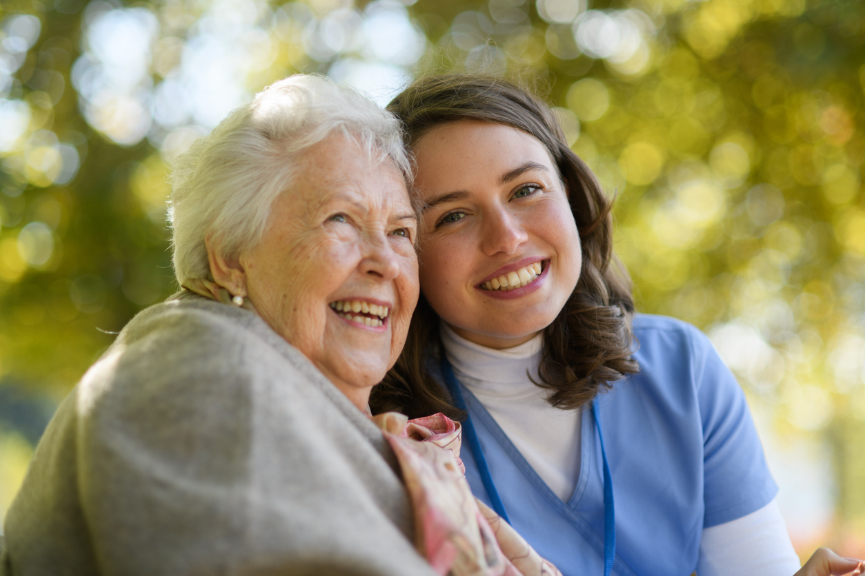 A happy young female caregiver and a joyful senior woman sit close together outdoors, smiling at the camera and sharing a warm, caring moment.