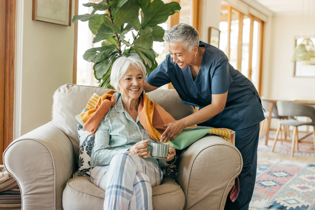 A caregiver adjusts a colorful blanket around an elderly woman sitting comfortably with a mug in a cozy living room setting.