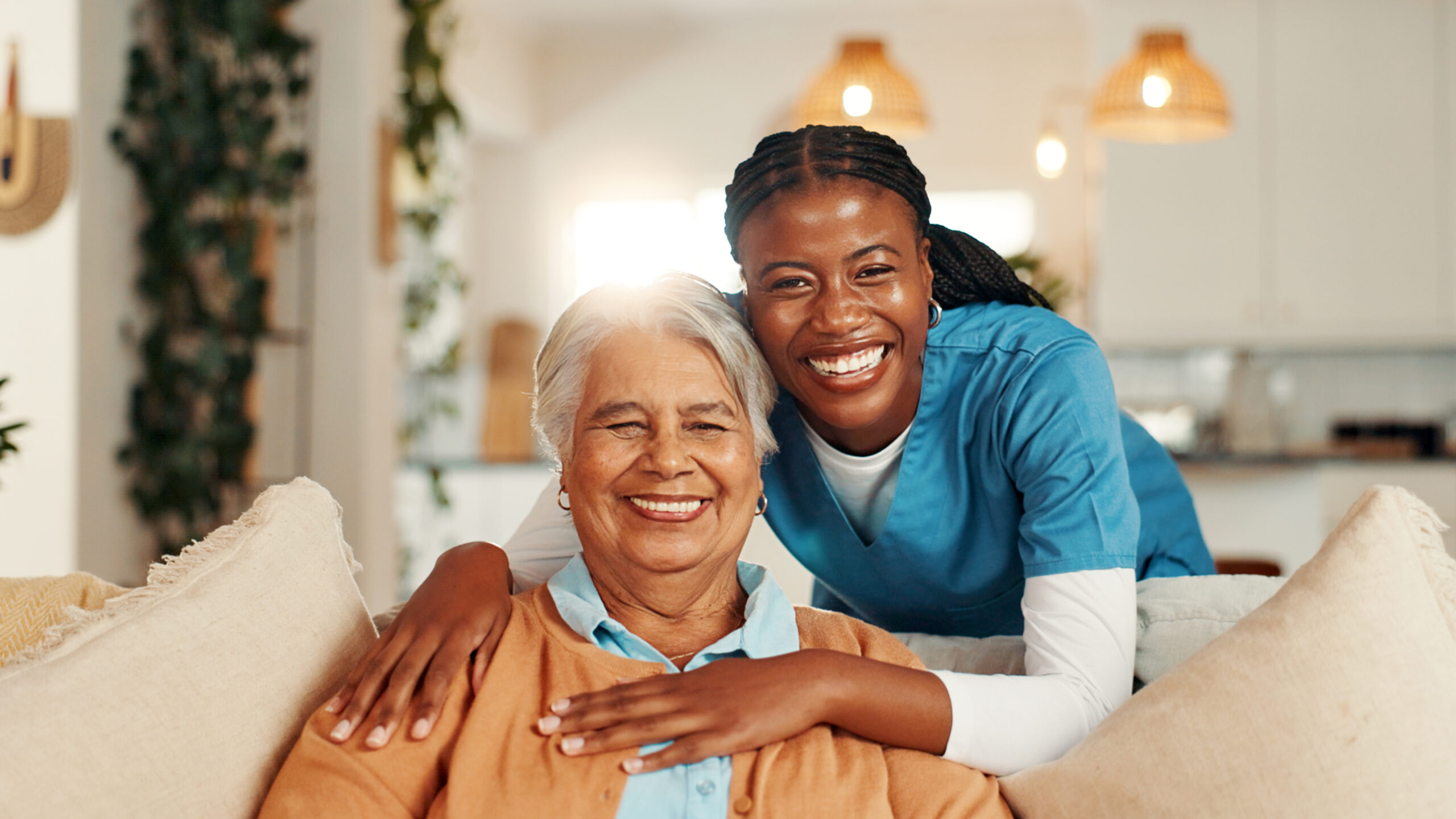 A caregiver and an elderly woman share a joyful moment, smiling while seated on a couch in a warmly decorated living space.