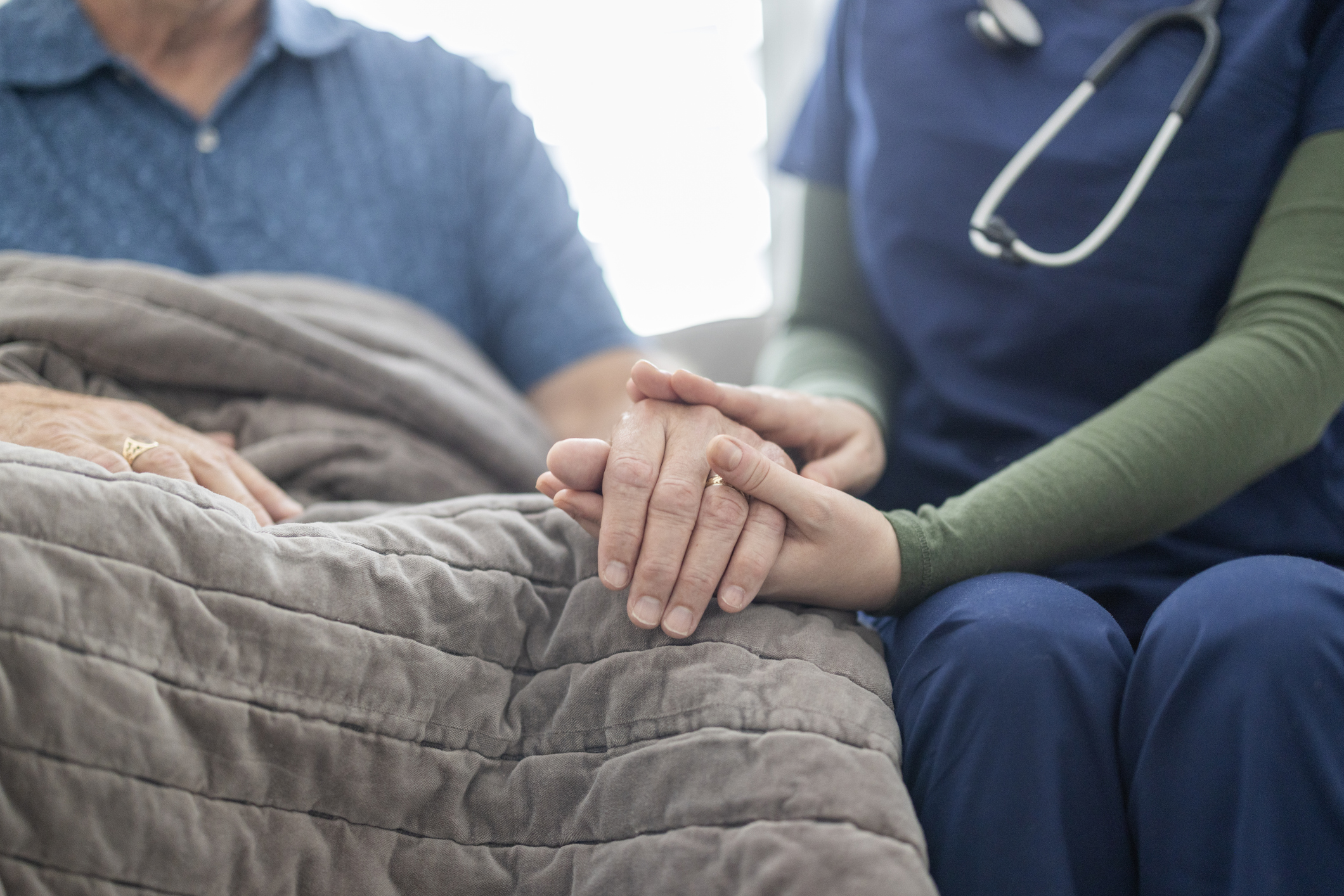 A caregiver gently holds the hand of a patient, both seated beside a soft gray blanket, communicating compassion.