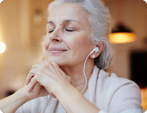 Smiling older woman wearing earphones, eyes closed, with hands clasped in a warm, softly lit indoor setting.