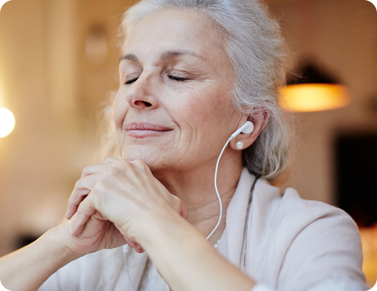 Smiling older woman wearing earphones, eyes closed, with hands clasped in a warm, softly lit indoor setting.