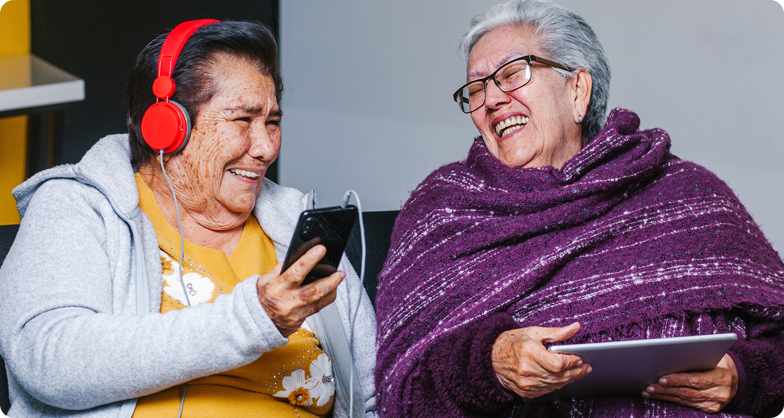Two elderly women share a laugh while using a smartphone and tablet, one wearing red headphones and the other a purple shawl.