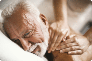Senior man receiving a massage, showing relaxation and care, while a therapist's hands are gently placed on his shoulder.