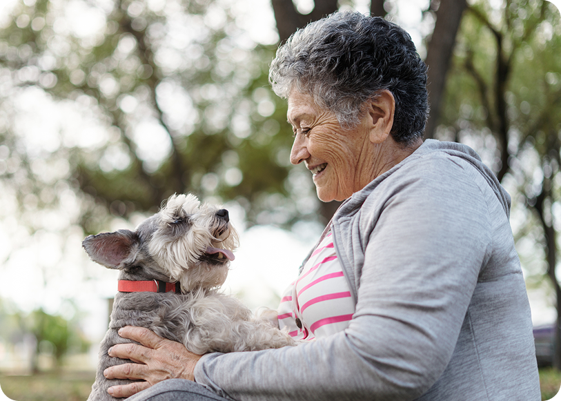 An older woman joyfully interacts with a small dog in a park, surrounded by trees and natural light.