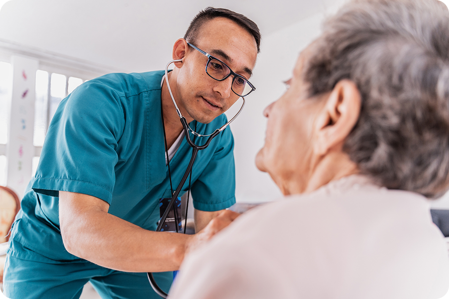 A healthcare professional in a teal uniform examines an elderly patient with a stethoscope in a bright consultation room.
