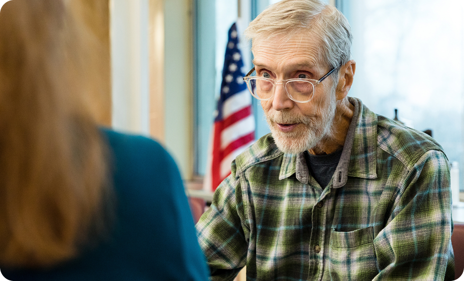 An older man with glasses, wearing a plaid shirt, engages in conversation with a person seated nearby.