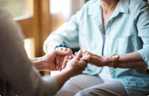 An elderly woman receives supportive touch therapy from a caregiver, both seated with hands gently clasped together.