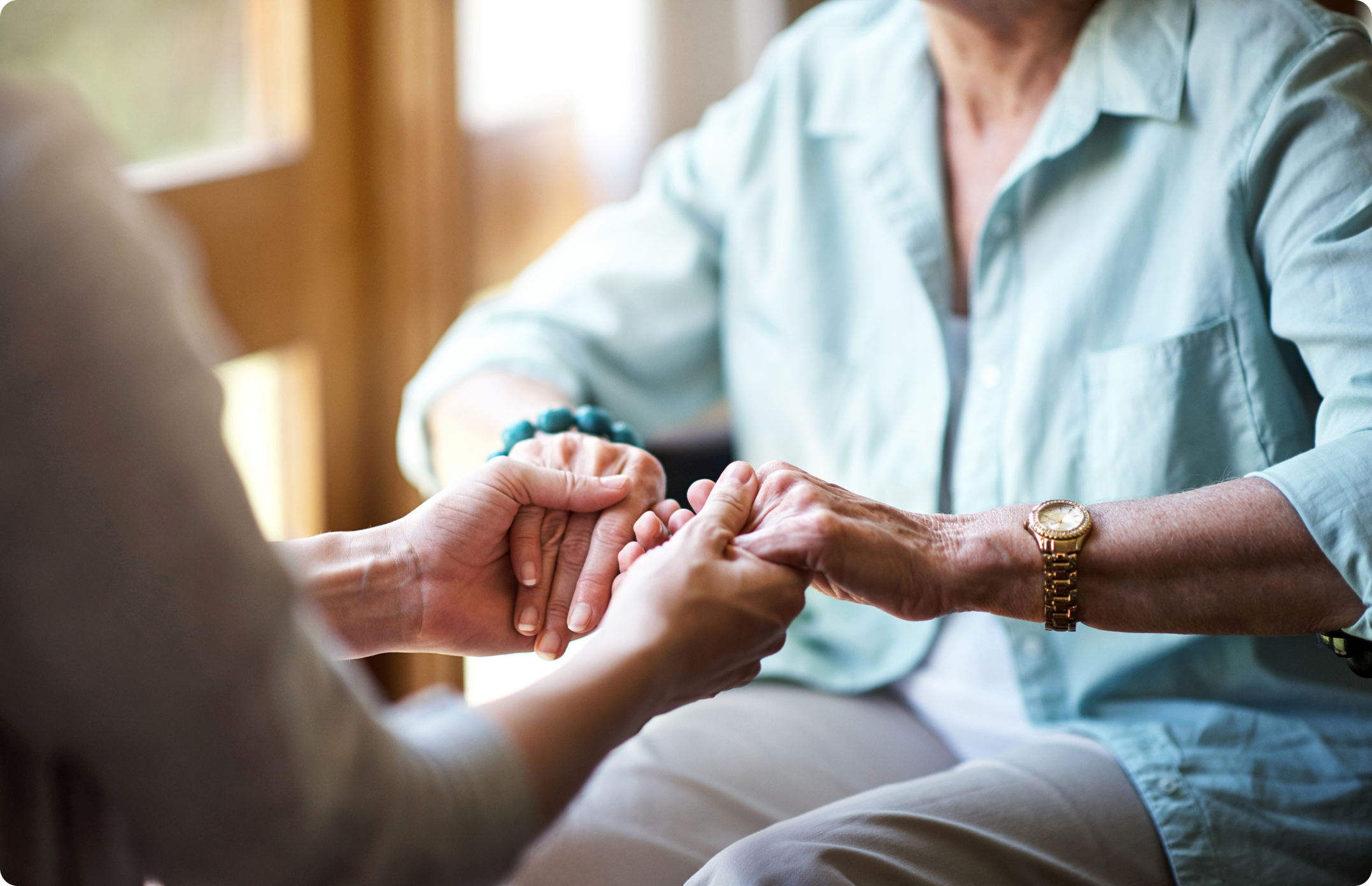 An elderly woman receives supportive touch therapy from a caregiver, both seated with hands gently clasped together.