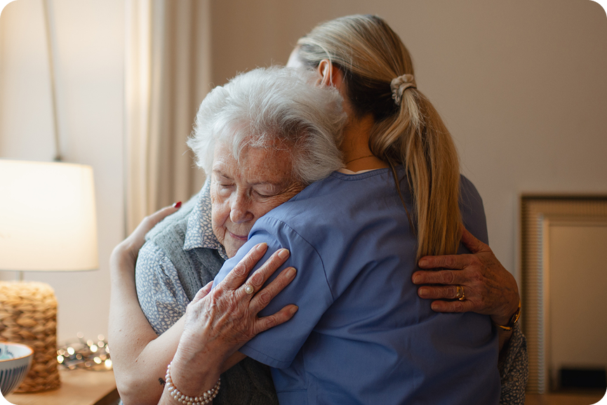 A compassionate caregiver in blue scrubs embraces a comforted elderly woman, as they share a warm hug.