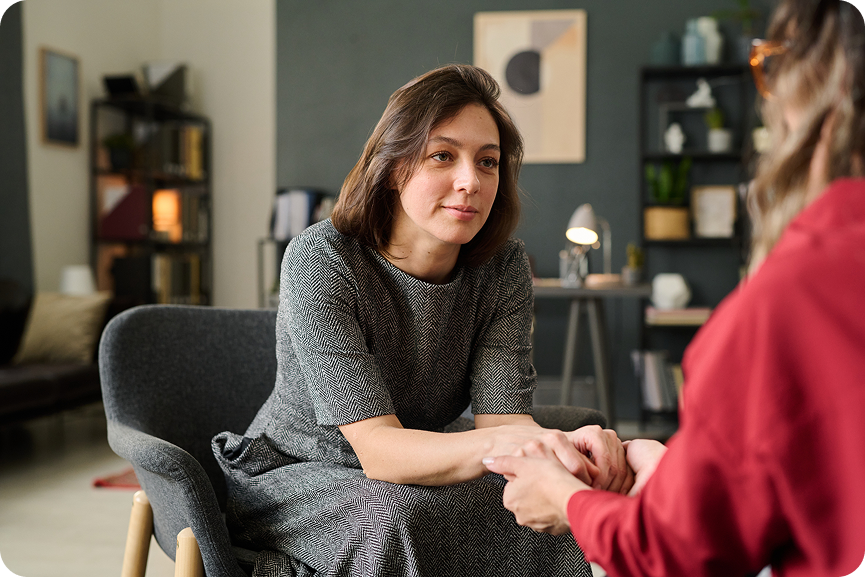 A concerned woman sits in a chair, holding the hands of a person sitting across from her, sharing a serious moment in a living room.