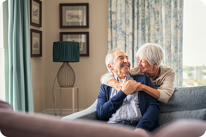 A joyful senior couple embraces on a couch. The woman stands behind the man with her arms around his neck, as they both smile warmly at each other in a cozy living room.