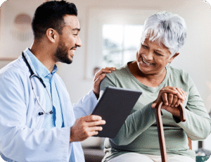 A friendly male doctor in a white coat shows a smiling senior woman a digital tablet, with his hand on her shoulder as she leans on a cane.