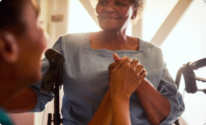 A smiling senior woman in a wheelchair holds the hands of a caregiver, with a warm and comforting look on her face.
