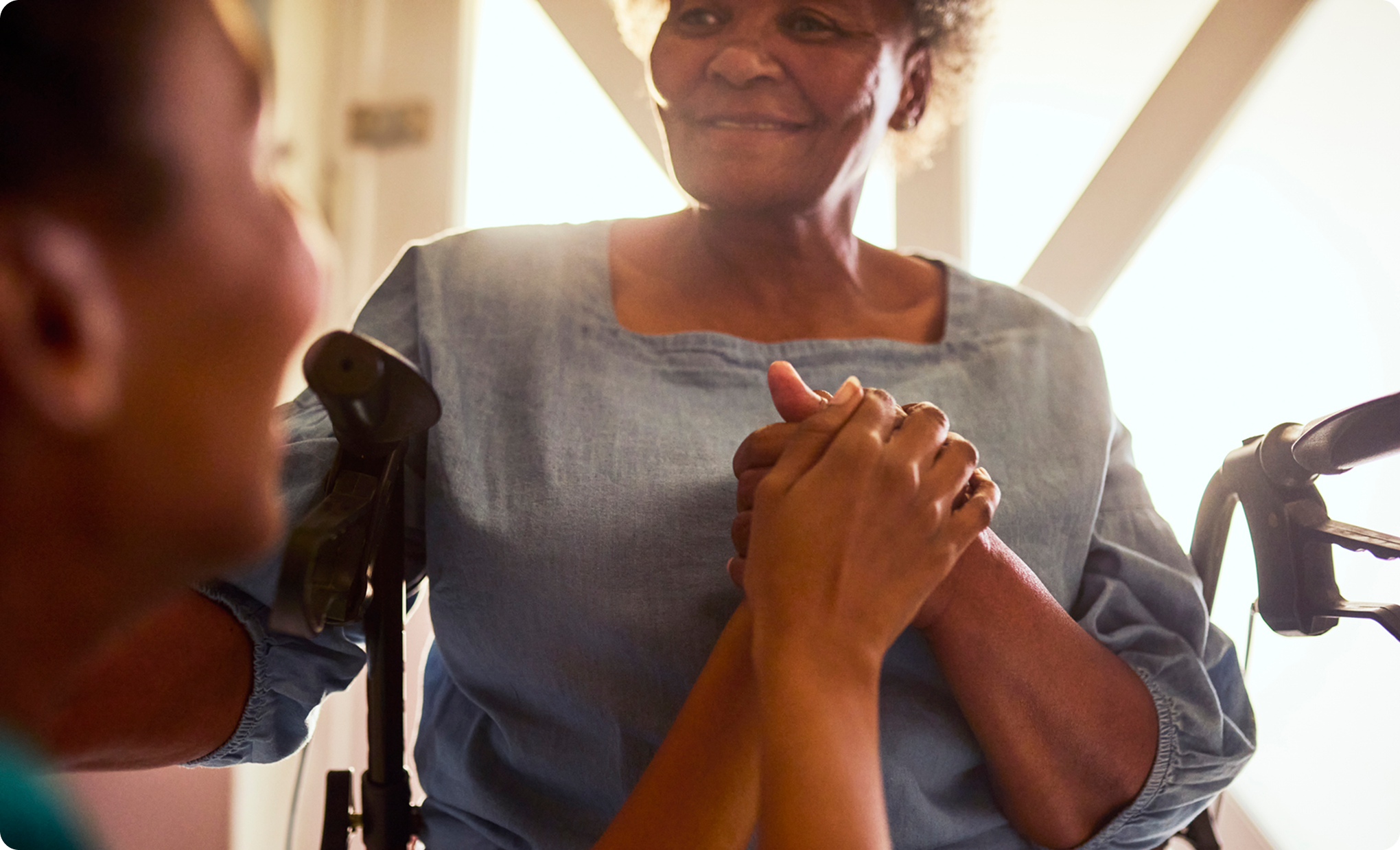 A smiling senior woman in a wheelchair holds the hands of a caregiver, with a warm and comforting look on her face.