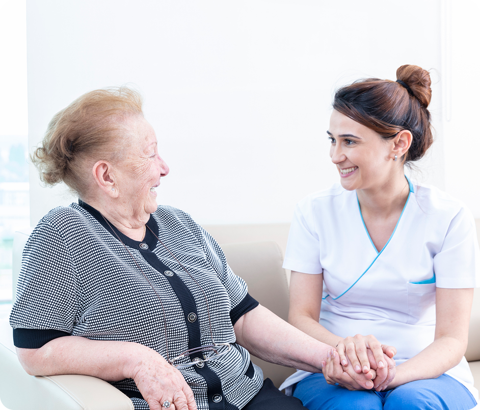 A female caregiver holds a senior woman's hand as they smile and talk together.
