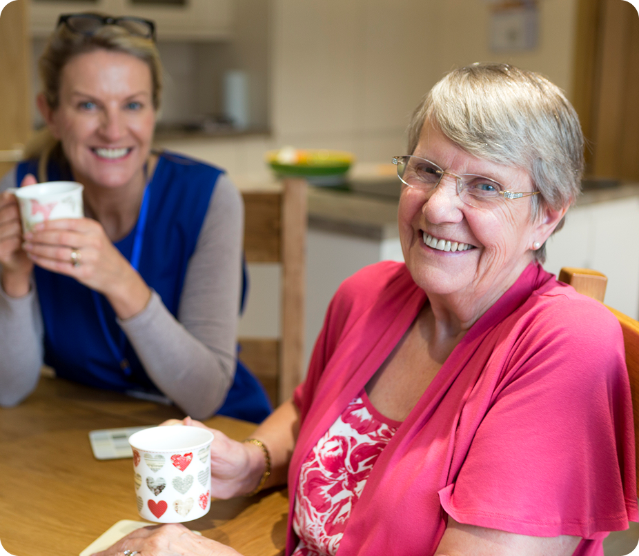 A friendly female caregiver and a senior woman smile at the camera while holding mugs at a kitchen table.