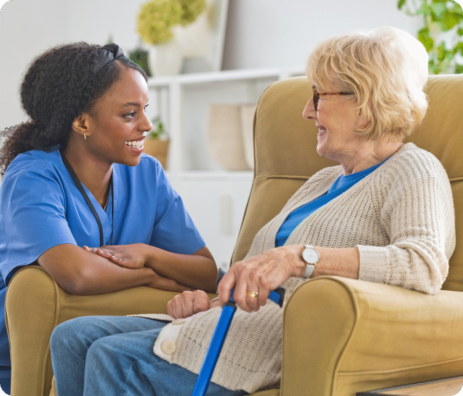 A Black female caregiver in blue scrubs smiles while sitting with a senior woman in a chair.