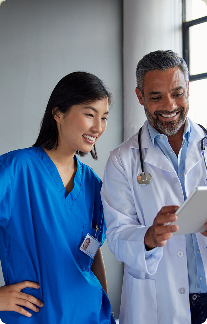 A male doctor and a female nurse, both smiling, look at a digital tablet together while standing in a hospital hallway.