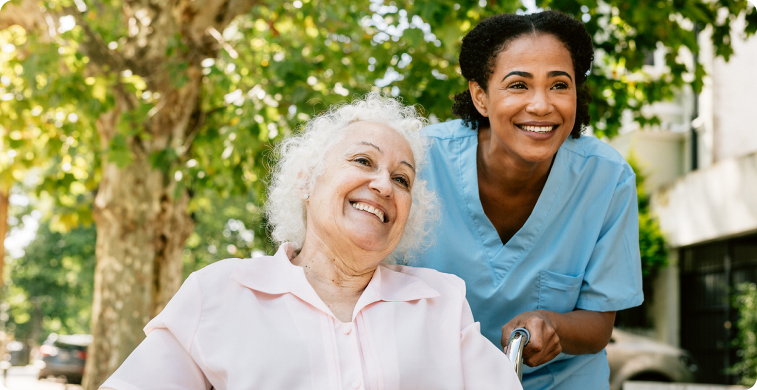 A caregiver in scrubs smiles with her arm around a senior woman outdoors.