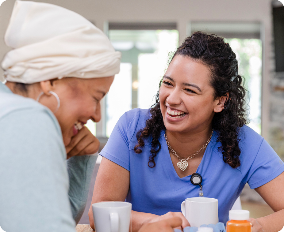 A caregiver laughs with a senior woman while sitting at a table with mugs and medication.