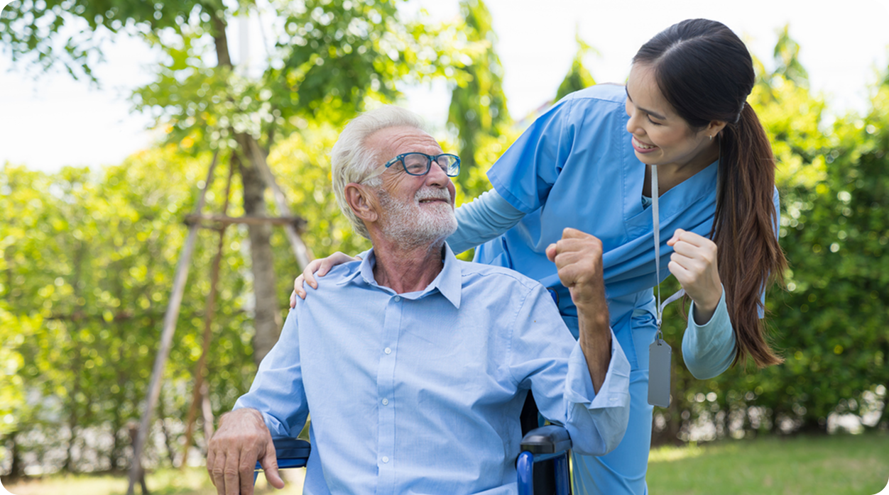 A female caregiver in blue scrubs smiles at a happy senior man in a wheelchair who is flexing his bicep.