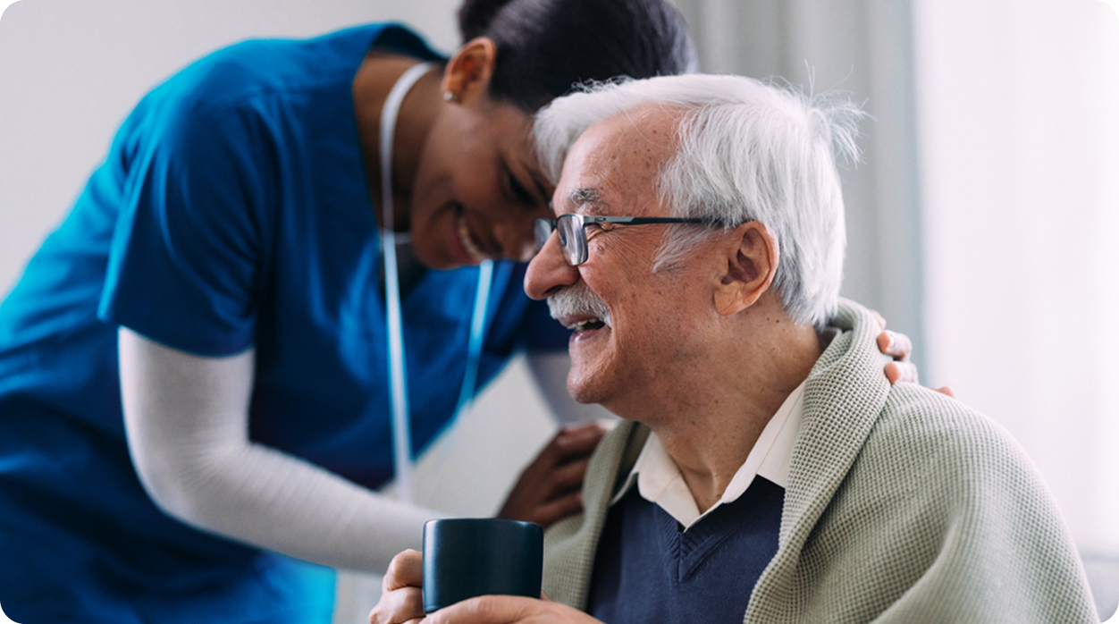 A female caregiver smiles and embraces a senior man as he laughs while holding a mug.