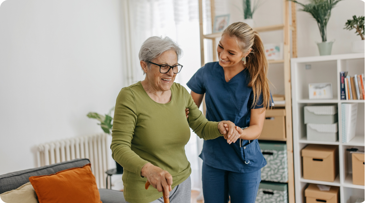 A smiling caregiver helps a senior woman with a cane walk across a living room.