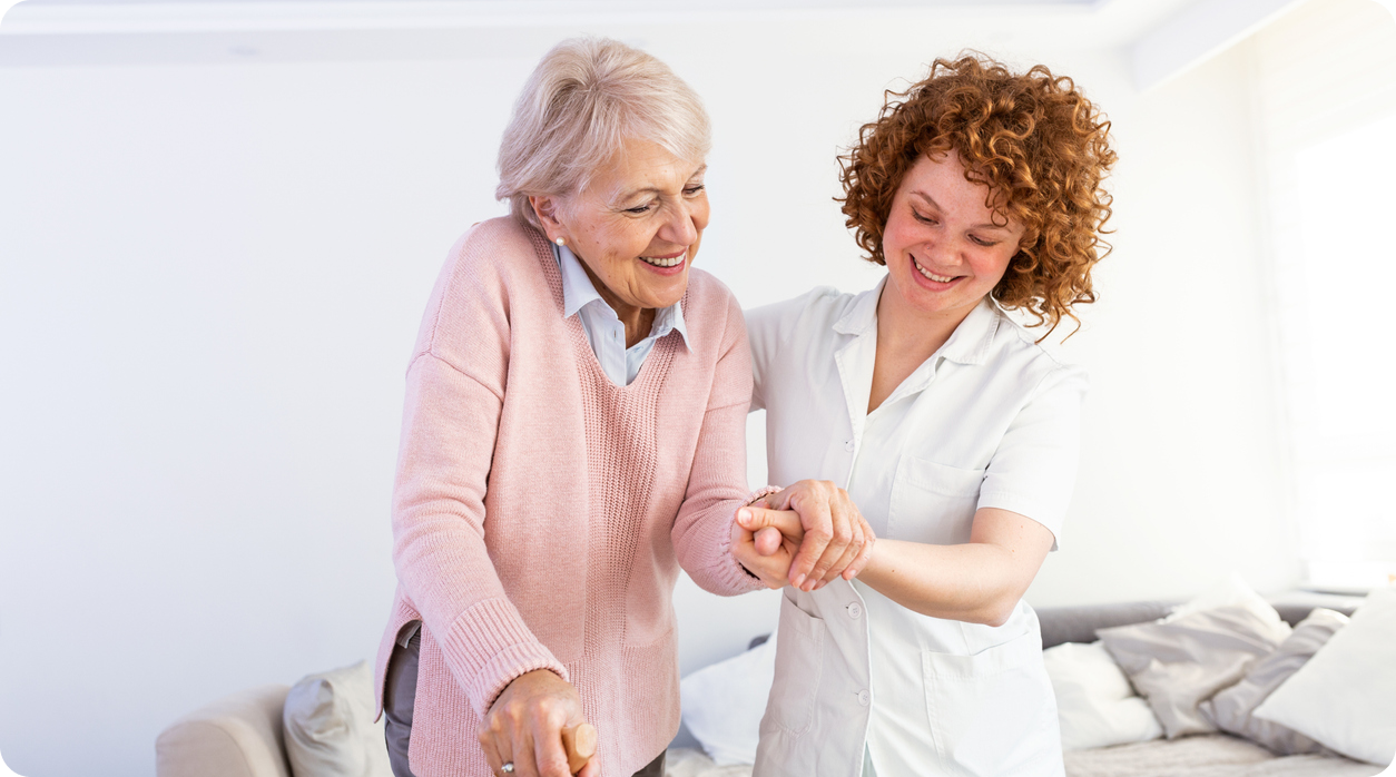 A smiling caregiver helps a senior woman with a walker, holding her hands for support.