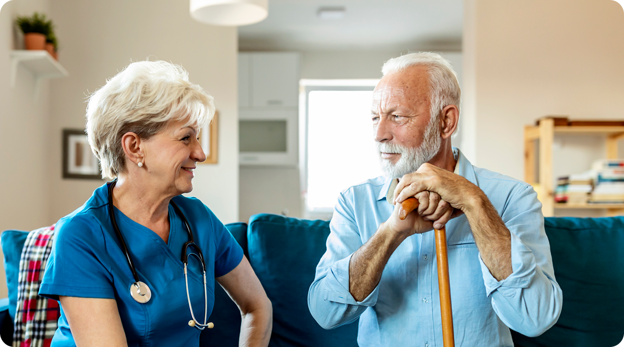A smiling female caregiver in blue scrubs talks to a senior man with a beard who is holding a walking stick.