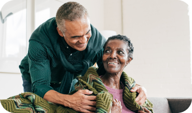 A smiling man stands behind a senior woman, gently wrapping a blanket around her shoulders and looking at her with a caring expression.