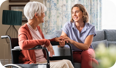 A friendly caregiver sits on a couch and holds the hands of a senior woman in a wheelchair.