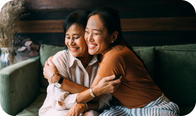 A happy younger woman hugs a laughing older woman from behind on a couch, sharing a joyful, loving moment.