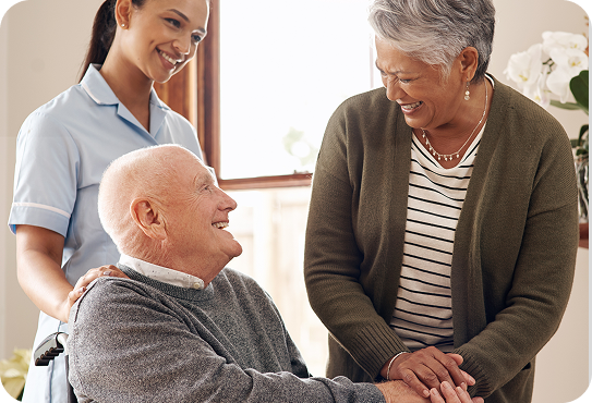 Smiling elderly man converses with two women, one in a caregiver uniform, in a warm, home-like environment.