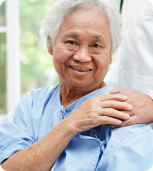 Smiling elderly woman in a blue medical gown sits comfortably, supported by a healthcare professional's hand on her shoulder.