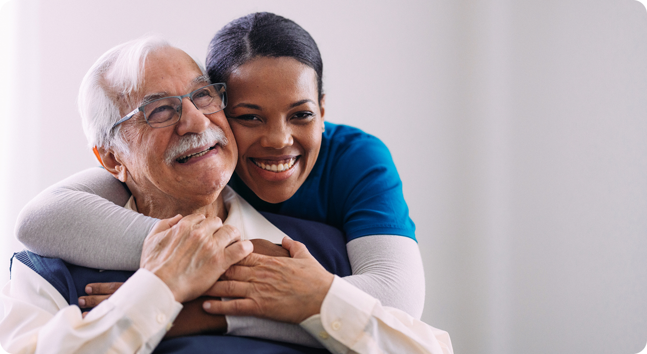 A caregiver embraces an elderly man, both smiling warmly in a bright, comfortable indoor setting.