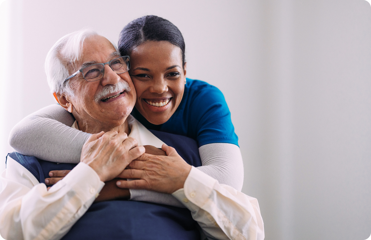 A caregiver embraces an elderly man, both smiling warmly in a bright, comfortable indoor setting.
