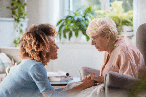 A caregiver interacts with an elderly woman, holding hands in a bright, plant-filled room promoting comfort and connection.